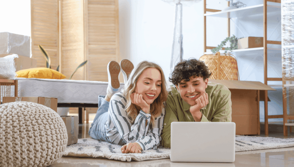 Paar liegt in neuer Wohnung auf dem Boden und informiert sich am Laptop, umgeben von Umzugskartons – Thema Warmmiete, Mietkosten und Wohnungseinzug.