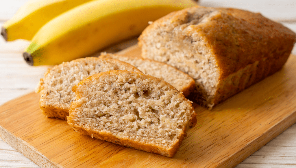 Saftiges Bananenbrot in Scheiben auf einem Holzbrett mit frischen Bananen im Hintergrund.